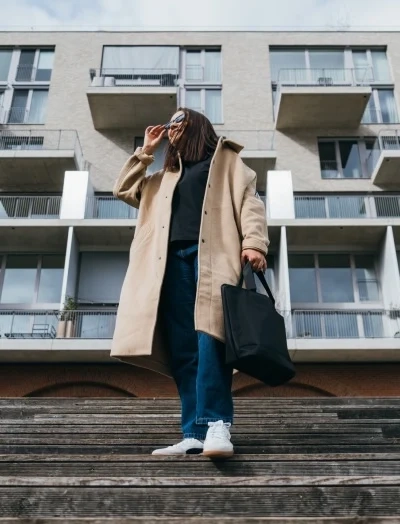 Mlada punca oblečena v svetlem plašču, s torbo v rokah in sončnimi očali, stoji na stopnicah pred blokom.i